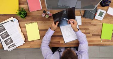 Hastanın eklem röntgenini inceleyen odaklanmış doktor tıbbi tarih ve üst görüş üzerine notlar alıyor. Klinikteki masada insan kemiğinin röntgenine bakan erkek ortopedi uzmanı.