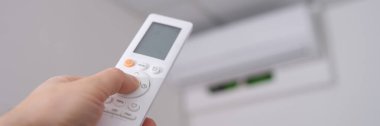 Woman hand holds remote control pointing at air conditioner in room. Adjusted to ambient temperature