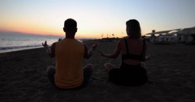 Couple yoga for meditation and meditating at serene sunset on beach. Girl and man are relaxing in lotus position in calm zen moment