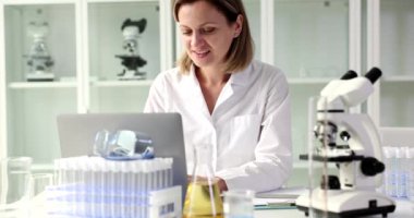 Smiling woman scientist or student using laptop computer and microscope. Online training and chemical data in lab