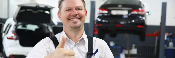 車のサービスを背景に親指で制服を着た若いハンサムな男の肖像画 推奨品質の車の修理コンセプト — ストック写真