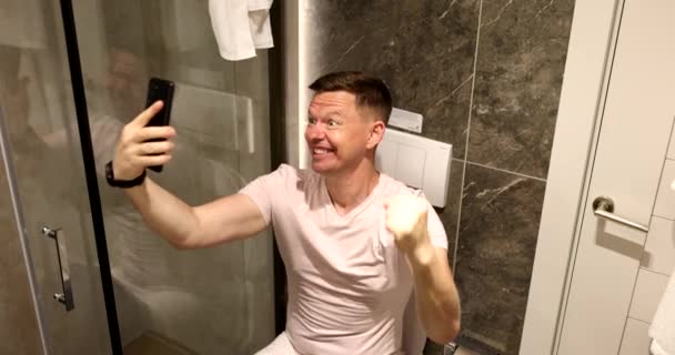 幸せな男は自宅でトイレに座って携帯電話を使用しています 素晴らしいニュース成功とガジェットへの中毒 — ストック動画