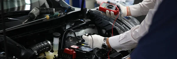 Professional auto mechanic works in a car service and tests operation of engine with tester. Mechanical service using multimeter to check voltage level in car battery