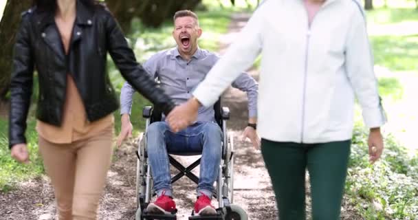 2人の若い女性が車椅子4K映画の中に座って不満男から離れて歩く 障害の概念を持つ人々から愛する人の拒否 — ストック動画
