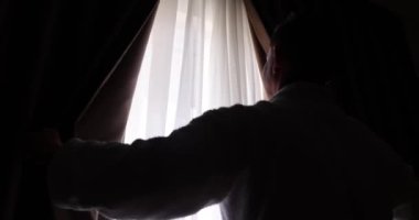 Adam dışarıdaki manzaraya bakmak için koyu perdeleri açar. Yeni bir günün ve daha parlak bir geleceğin başlangıcı