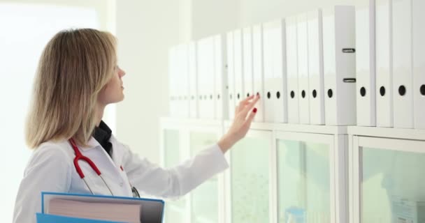 医者は棚から輪で白いフォルダを取ります 診療室で資料を探す制服姿の女性 — ストック動画