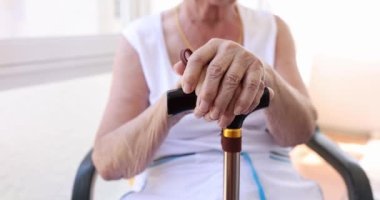 Sandalyede oturan bir emekli bastona yaslanır, yakın plan, yüzeysel odaklanma. Kırışık eller, bakımevi.