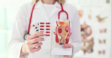 Kadın doktor elinde patoloji olan rektum modelini tutan hastaya su toplayan ilaçlar veriyor. Beyaz önlüklü doktor klinikte tedavi öneriyor.