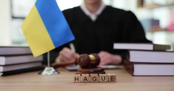 木製の立方体で作られた単語ハーグは ウクライナでの戦争中の犯罪に関する文書にメモを取るローブの女性弁護士に対して嘘をつく 裁判所のテーブルの上の小さなウクライナの旗 — ストック動画