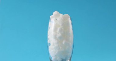 Beyaz şeker dolu şeffaf bardak parlak mavi stüdyo arka planında duruyor. Şekerin büyük miktarlarda zararlı kullanımı kavramı. Diyabet Hastalığı