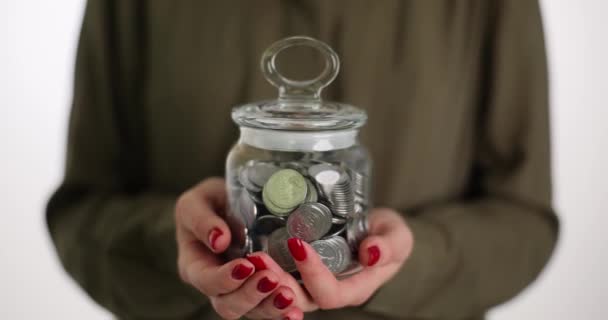 女性は銀のコインでいっぱいの巨大な透明な瓶を持っています 光の背景にコインの貯蓄を持つ女性の人 スマート 支出の概念とホーム バンク — ストック動画
