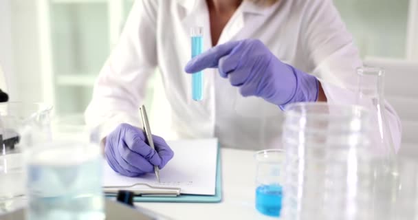 科学者は 研究の結果を書き留める青い試薬で試験管を保持しています 磁気攪拌機付き滅菌テーブルの研究室の労働者とブランク付きのプランケット — ストック動画