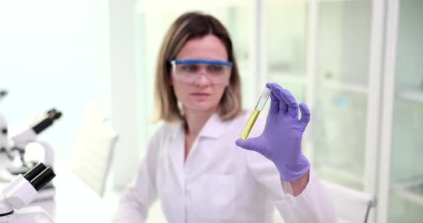濃縮女性研究者は 顕微鏡下での検査のために黄色の液体のデスク保持管に座っています 女性でホワイト研究室コート実験遅い動き — ストック動画