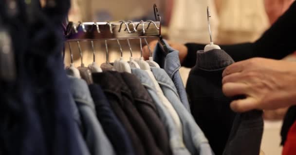 男は店内のコートでいっぱいのスタンドの前に立っている間 黒と青のデニムジャケットの間を選択します 男はジャケットを買うために選択します — ストック動画
