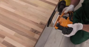 Male employee in work uniform processes parquet board with grinding machine at workplace in room of apartment. Specialist uses device on plain wooden surface slow motion