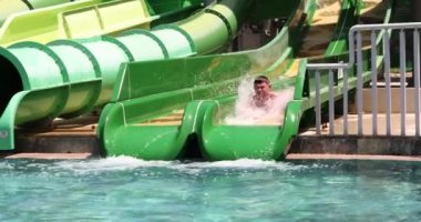 Cheerful male person in swimming trunks descends from water slide into pool in water park. Entertainment and recreation concept at resort slow motion