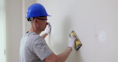Builder in helmet and goggles polishes plaster spot on wall in restored room. Employee fixes holes and trenches on drywall panel with tool
