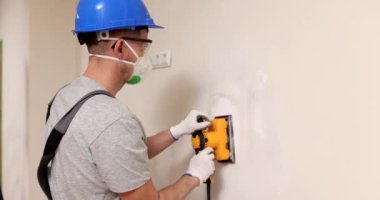 Labourer in helmet with respirator polishes plaster coverage on wall in room. Builder fixes holes on drywall panel with electrical grinder tool