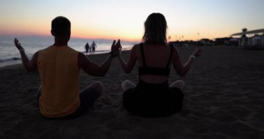 Gün batımında, bir adam ve bir kadın kumların üzerinde oturmuş meditasyon yapıyorlar, yakın plan. Sahilde yoga konsepti