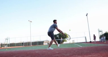 Bir erkek ve bir kadın açık tenis kortu oynuyorlar, yavaş çekimde. Popüler bir spor oyunu öğretmeyi saymazsak, oyuncular