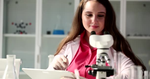 医学検査官は顕微鏡を検査し 実験室での実験や研究に関する情報を記録する 結果の治療とドキュメンテーションを探している微生物学の女性 — ストック動画