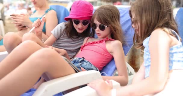 三个带着平板电脑的好奇女孩看了一部供孩子们在海滨度暑假的电影 孩子们躺在沙滩上的日光浴中研究孩子们的申请 — 图库视频影像