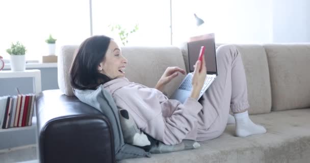 Woman Lying Sofa Laptop Takes Video Call Smartphone Joyful Greeting — Vídeo de Stock