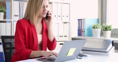 Kırmızı iş ceketli genç bir kadın telefonla konuşuyor ve bilgisayar klavyesinde 8K yazıyor. İş konseptinde müşterilerle uzaktan çalışma