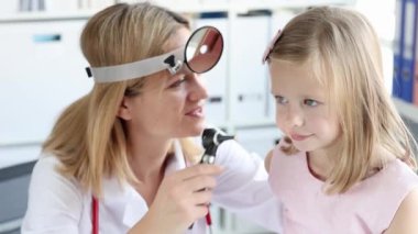 Ön reflektörlü Otorhinolaringolog doktor klinik 4K filminde otoskoplu küçük kız kulağını inceliyor. Çocuk konseptinde akut otitis medyasının teşhisi ve tedavisi