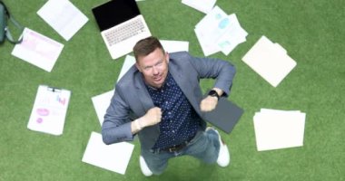 Businessman happily dances on green lawn, lies laptop and business charts. Building a successful career concept