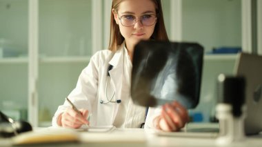 Konsantre kadın cerrah travmatolojist röntgenlere bakar kırık ya da florografiyi inceler. Doktor, teşhis ve tıbbi bakım kavramının sorunlu olduğunu düşünüyor.