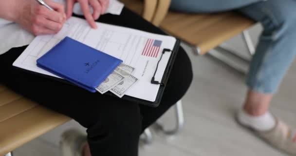 人々はライトルームでビザ申請書に記入します 女性は パスポートとドル紙幣で椅子に座っている文書に署名する — ストック動画