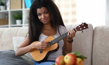 Kahverengi ukulele gitarla çalan gülümseyen Afro-Amerikan kadın portresi. Latin müzisyen rahat koltukta oturuyor. Neşeli bayan kameraya mutlulukla bakıyor. Müzik ve eğlence konsepti