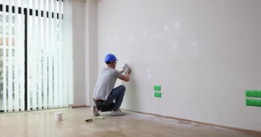 Koruyucu miğferli tamirci restore odasındaki beyaz duvarı parlatıyor. İşçi, yeni dairede macunların bozulmasından sıyrıldı