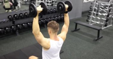 Sportif görünümlü genç adam dambılları başınızın üstüne bastırıyor. Spor salonunda çalışan kişi kol ve sırt kaslarının gücünü yavaş çekimde arttırır.