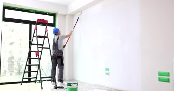 戴头盔的建筑者为新公寓刷墙壁 人在房间里涂上一层白漆 用滚子涂上长棒 室内翻修慢动作 — 图库视频影像
