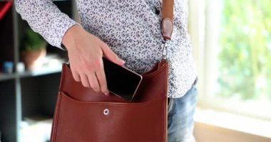 Woman puts smartphone in brown leather bag. Mobile data security in your phone