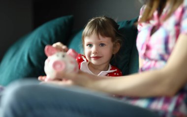 Anne ve kızı yeşil kanepeye oturup oynuyorlar. Kadın elinde pembe domuz kumbarası tutuyor. Neşeli çocuk para kutusuna para koydu..