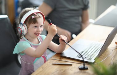 Çocuk kulaklıkla sandalyeye oturur ve mikrofonu elleriyle tutar. Kız bilgisayarda konuşuyor..