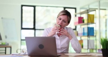 Aç kadın işçi, şirketteki masada dizüstü bilgisayara bakarak shawarma yiyor. İş yerinde öğle yemeği molası ve teknoloji konsepti ağır çekim