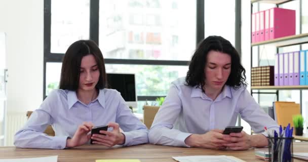 従業員のカップルは 会社のオフィスのテーブルに座っている携帯電話を使用しています 明るい前提で職場でのクライアントや技術とのチャットの概念 — ストック動画