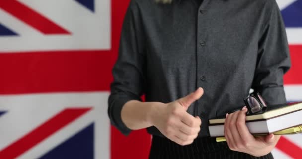 教科書とノートブックを持つ女性は 英語を勉強した後に親指を立てます イギリスの国旗の壁は英語学習の遅い動きに対する敬意を示しています — ストック動画