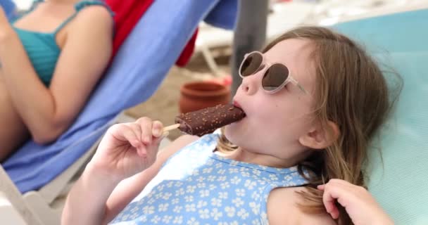 サングラスの小さな女の子は 母親の近くのサンラウンジャーに横たわるスティックにアイスクリームを食べます 家族と一緒にビーチで観光客や休暇 冷たい食べ物のスローモーションを食べる — ストック動画