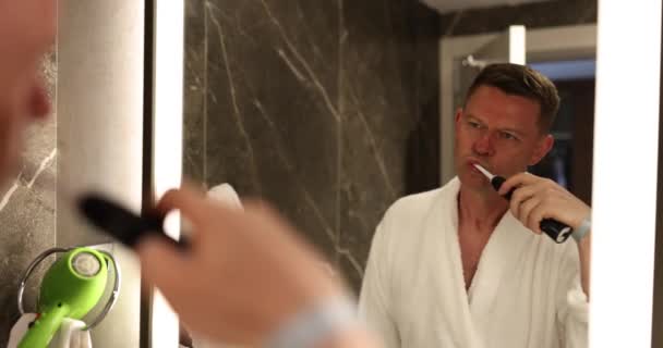 사람은 현대적인 욕실에서 백라이트 거울을 바라보는 칫솔로 치아를 닦습니다 일상적인 — 비디오