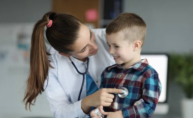 Doktor randevusundaki gülümseyen çocuğun yakın çekimi. Kalp atışlarını dinleyen ve çocuk kulaklarını kontrol eden profesyonel bir çocuk doktoru. Planlı check-up. Tıp ve sağlık hizmetleri kavramı