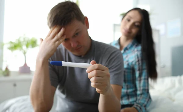 Man rubs his forehead looking at pregnancy test. Couple planned pregnancy together. Man is fully aware, there is reassessment values. Strange test result indicates onset an ectopic pregnancy