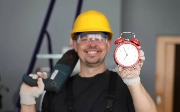Male builder helmet laughing showing alarm clock. Performance repair work within specified period. Noisy work with drilling holes concrete. Happy builder finished work. Deadlines and employee safety