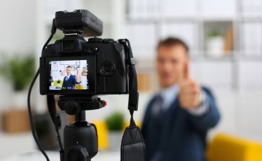 Erkek takım elbise ve kravat göstermek onaylamak işareti kol yapma promo videoblog veya fotoğraf oturumunda office video kamera tripod taşımak için. Vlogger tanıtım selfie çözüm ya da Finans Danışmanı yönetimi bilgileri