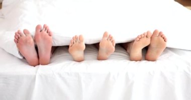 Üç kişi yorganın altında, bacakları dışarıda yatıyorlar. Üç çift ayak yatakta üçlü yaparken eğleniyor.
