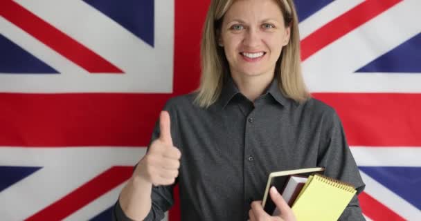 若い女性は言語学習のために親指をあきらめる イギリスの国旗の前に立っている笑顔の英語教師と学生が勉強することを奨励 — ストック動画